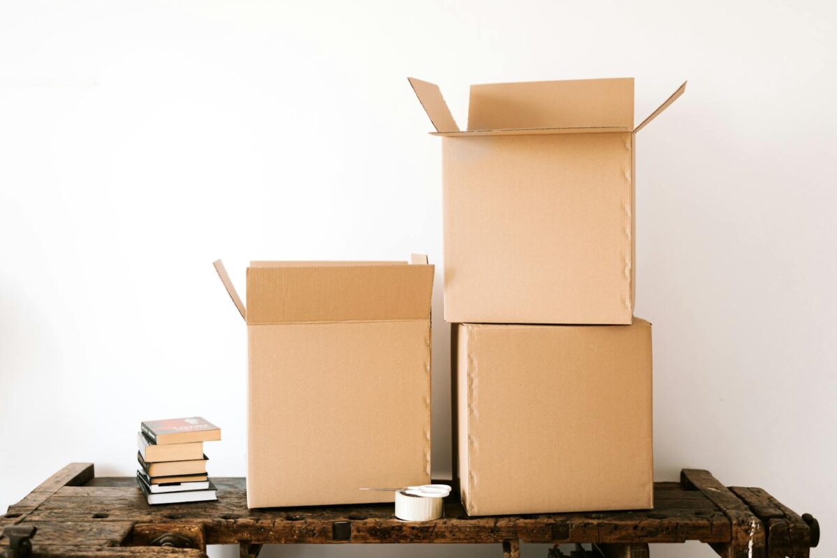 Opened cardboard boxes and stack of books placed on wooden shabby table near tape with scissors against white plain wall