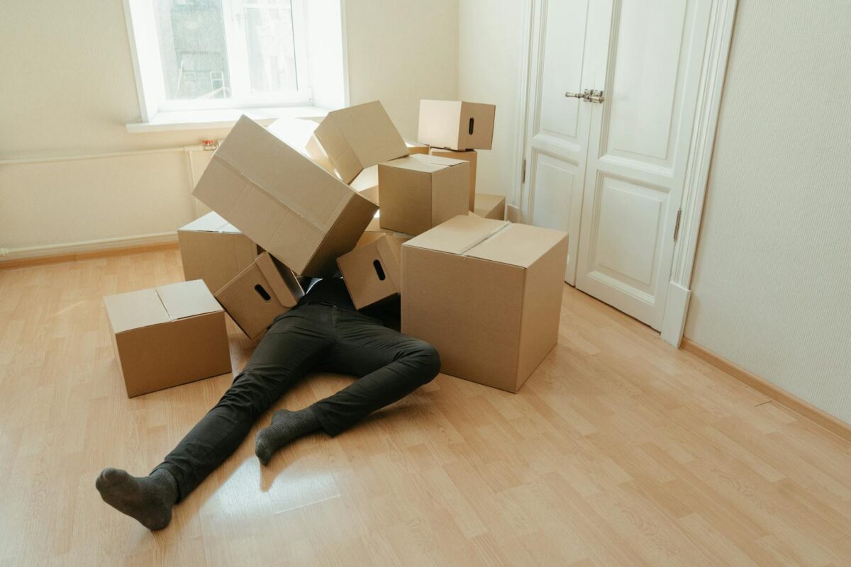 A person collapsed under stacks of cardboard boxes in an empty room, illustrating the stress of moving.