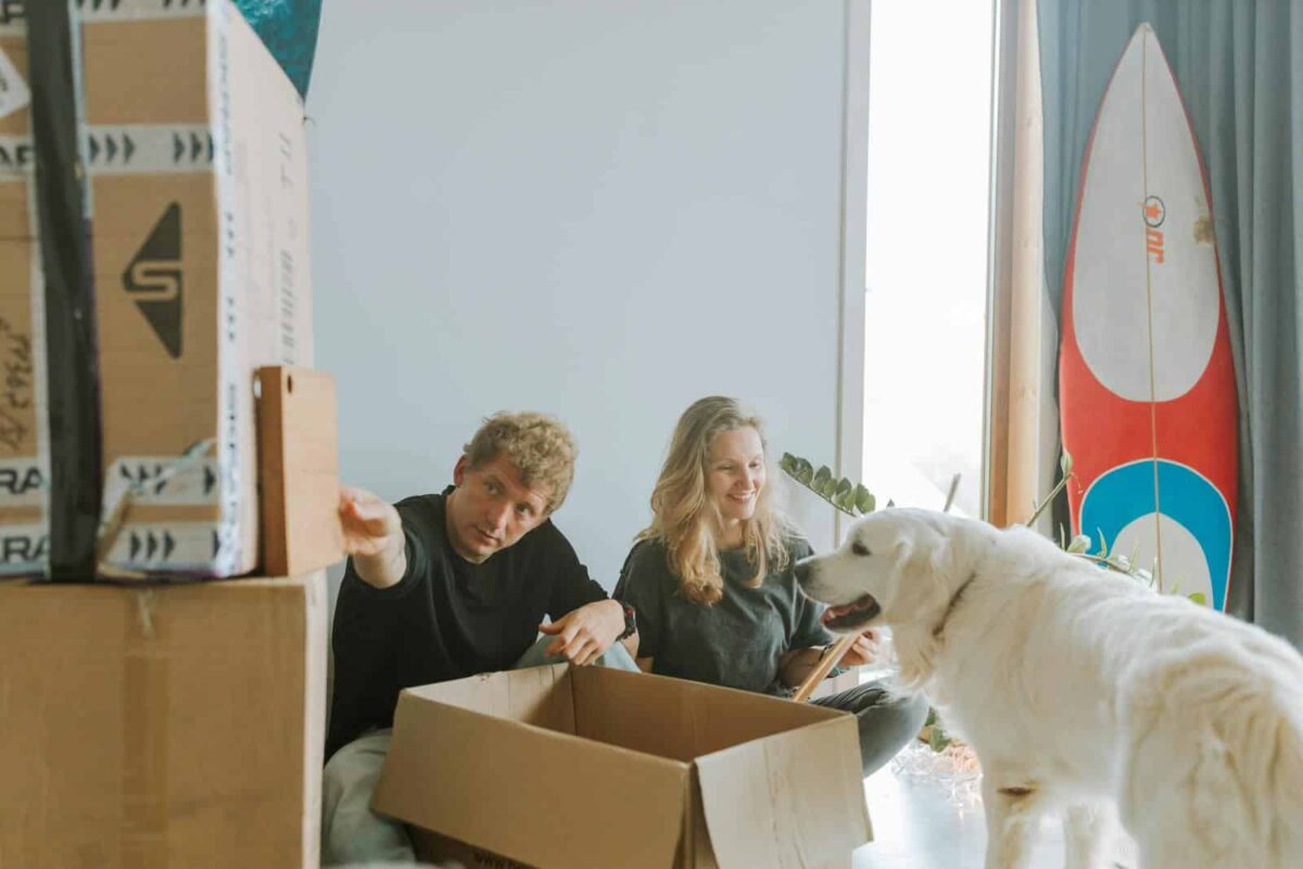 A cheerful couple unpacking boxes with their playful dog in a bright, modern living room.