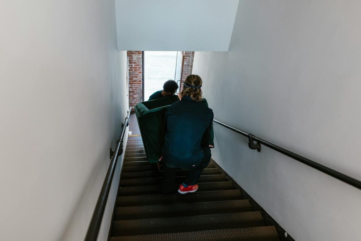 Two men carefully move a couch down a staircase in an indoor setting.